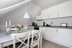 Ett modernt kök med vita skåp, ett matbord med stolar, en hängande taklampa och en kaffebryggare.