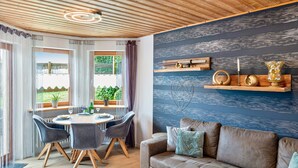 A modern living room with a wooden ceiling, a brown sofa, a dining table with chairs, and a view through large windows. - Holiday Apartment 'Quiet Basement Apartment with Terrace' with Private Terrace, Garden, and Wi-Fi (Altenkunstadt)