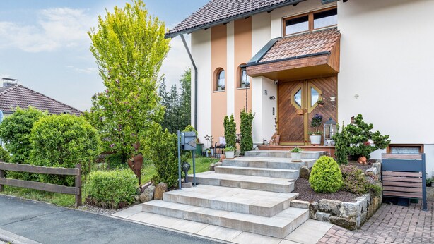 A two-story house with a stone staircase leading to the front door, surrounded by greenery and a wooden fence. - Holiday Apartment 'Quiet Basement Apartment with Terrace' with Private Terrace, Garden, and Wi-Fi (Altenkunstadt)
