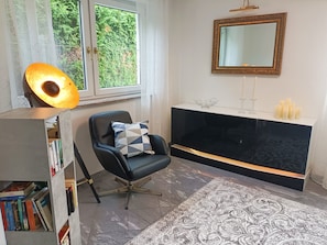 Une pièce comprenant un fauteuil en cuir noir, une bibliothèque, une cheminée avec un manteau de couleur blanche et une grande fenêtre donnant sur un paysage verdoyant.
