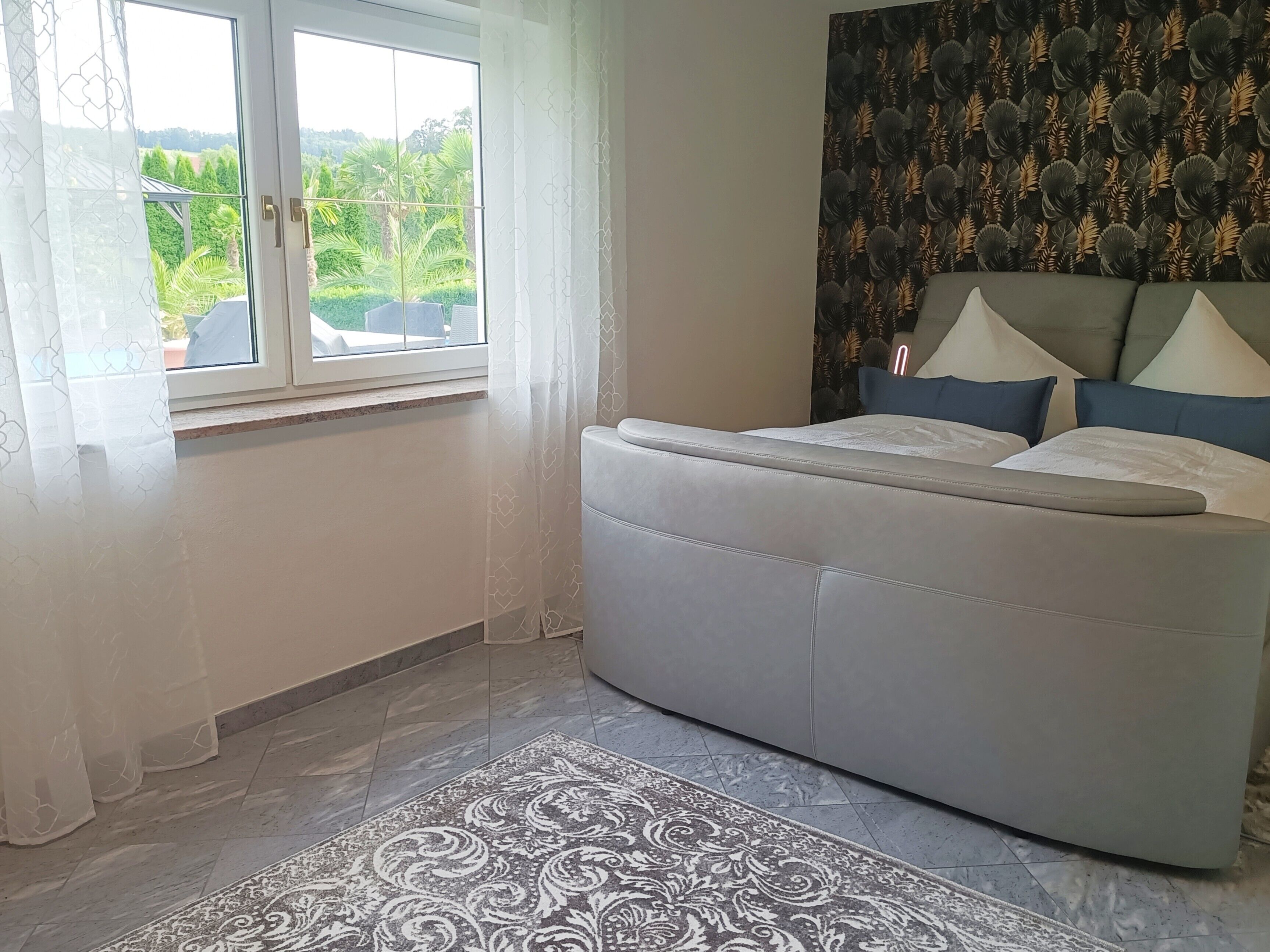 Ein Schlafzimmer mit einem Bett, einem Fenster mit Vorhängen, einem gemusterten Teppich und einer Wand mit Blättermotiv.