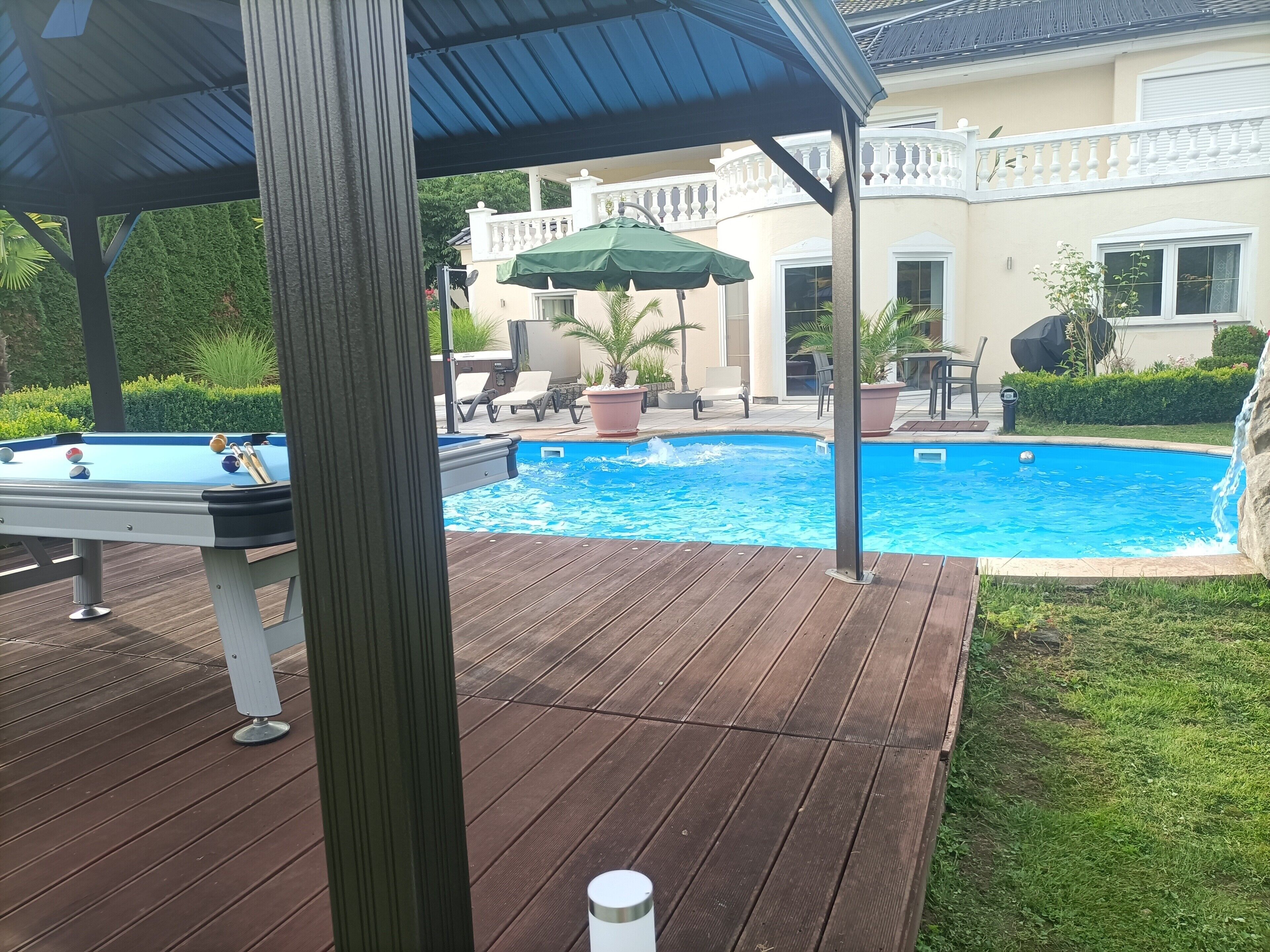 A swimming pool with a wooden deck, lounge chairs, and a covered area.