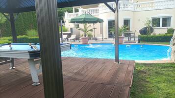 A swimming pool with a wooden deck, lounge chairs, and a covered area.