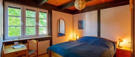A bedroom with a bed, a desk, a chair, a window, and a wooden ceiling.