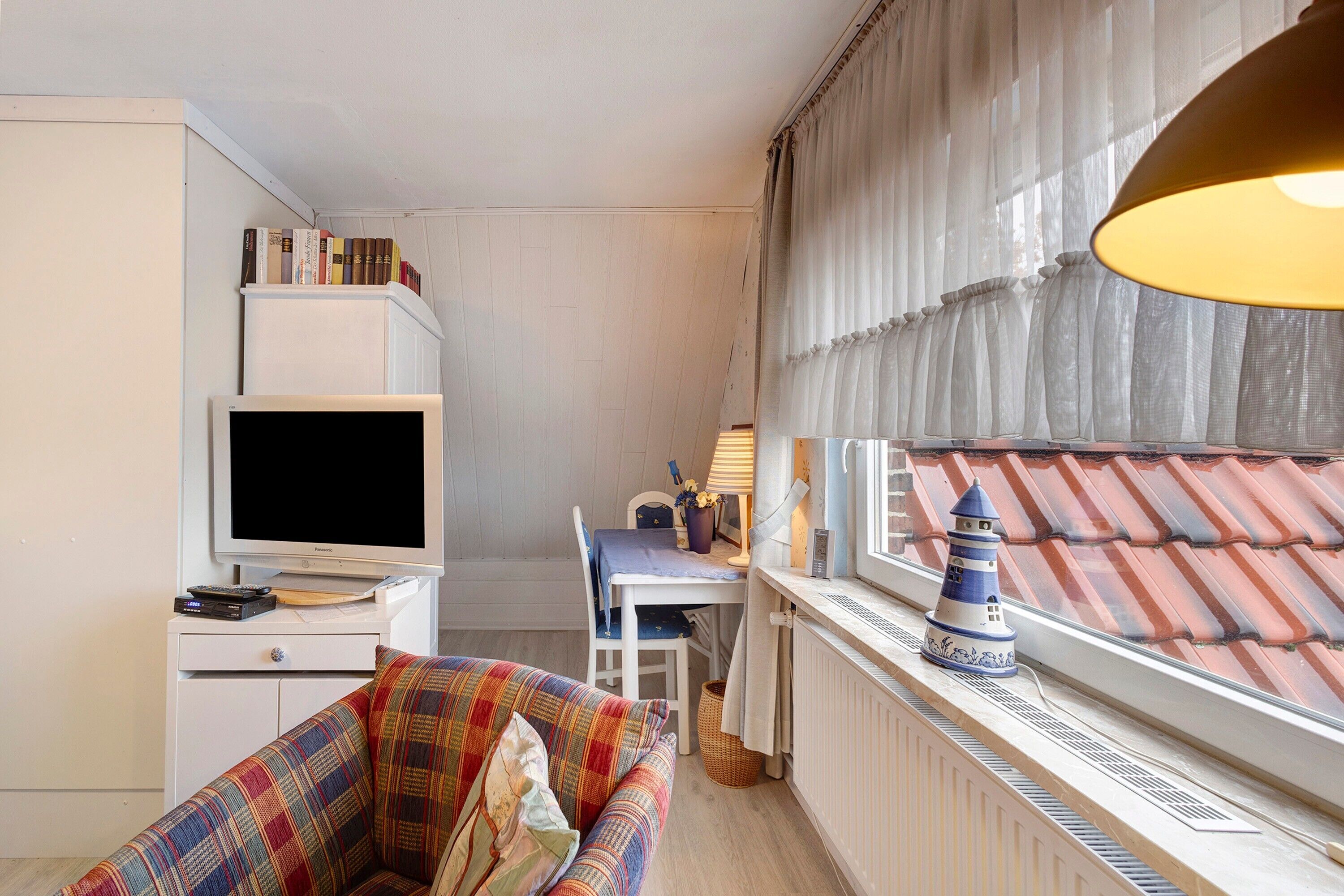 Una stanza compatta con televisione, un tavolino, una sedia, un divano, una finestra vista su un tetto in tegole e un modellino decorativo di un faro.