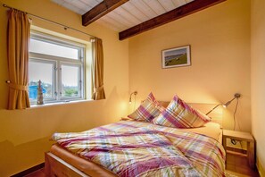 Un dormitorio con cama, cortinas, mesita de noche, lámpara y un cuadro en la pared.