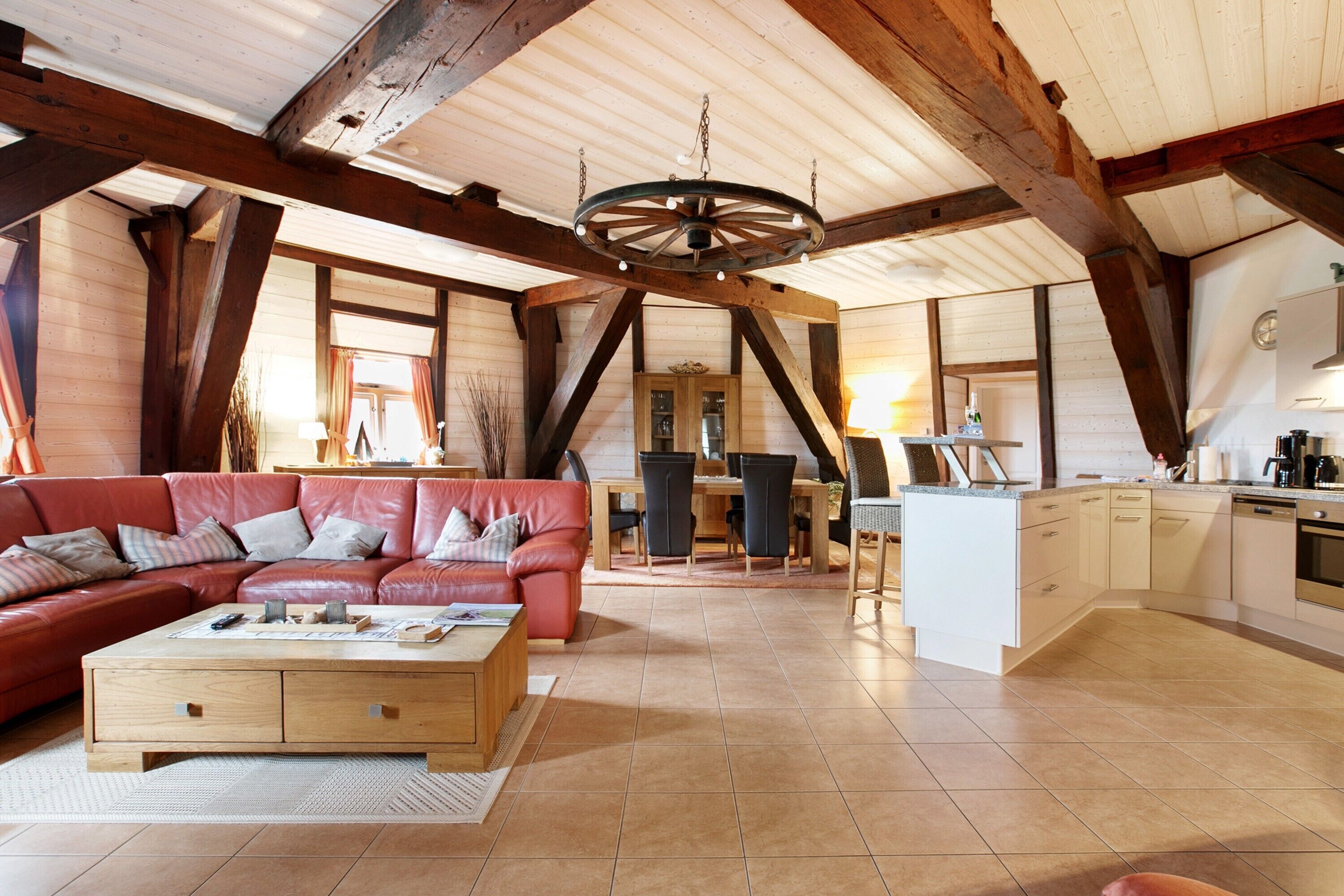 Ein geräumiger Loft mit hoher Decke, freiliegenden Holzbalken, einem roten Ledersofa, einem Holztisch und einer Küchenzeile mit weißen Schränken und einem Geschirrspüler.