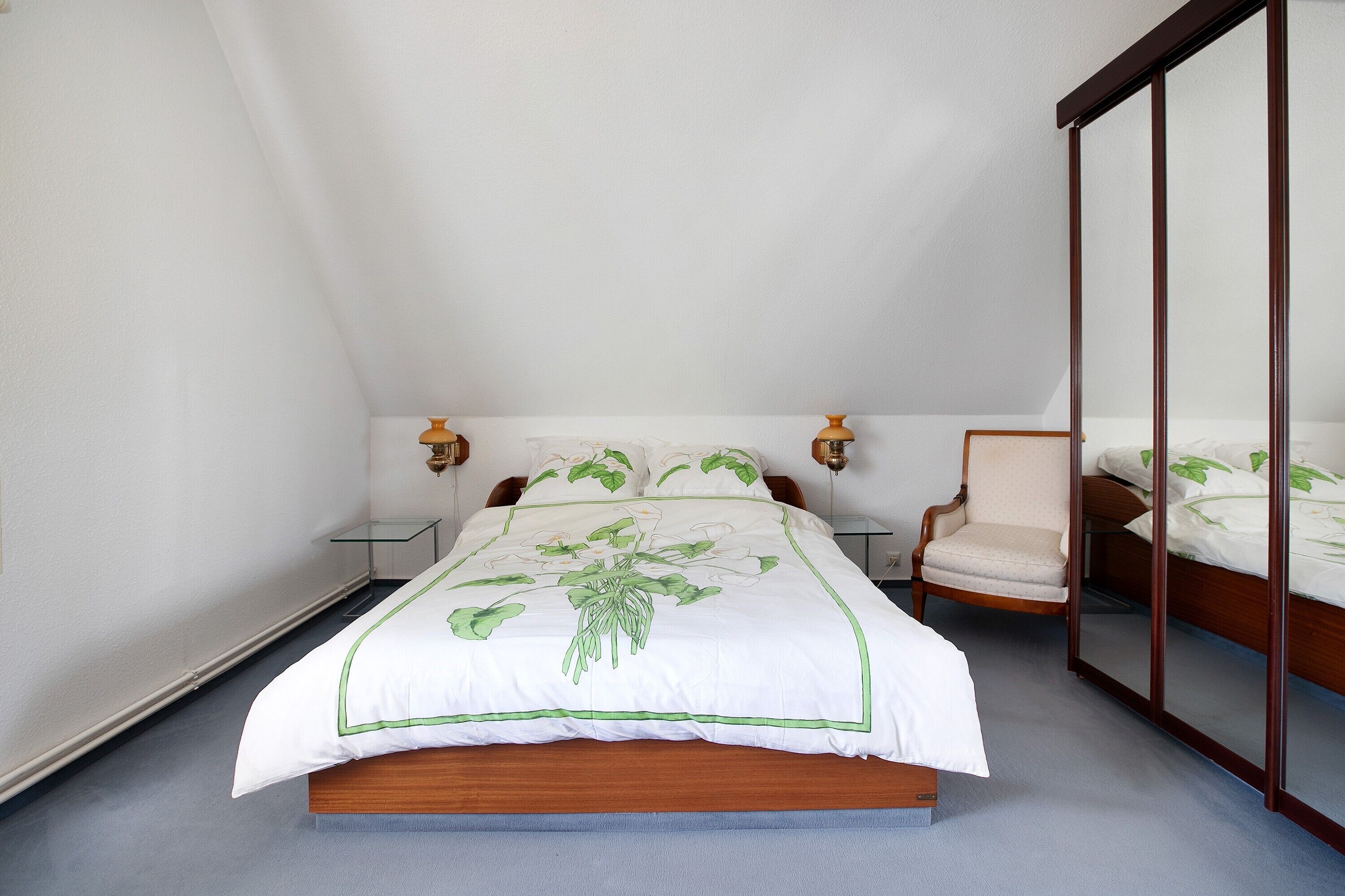 Een slaapkamer met een bed, stoel, nachtkastjes en een spiegel.