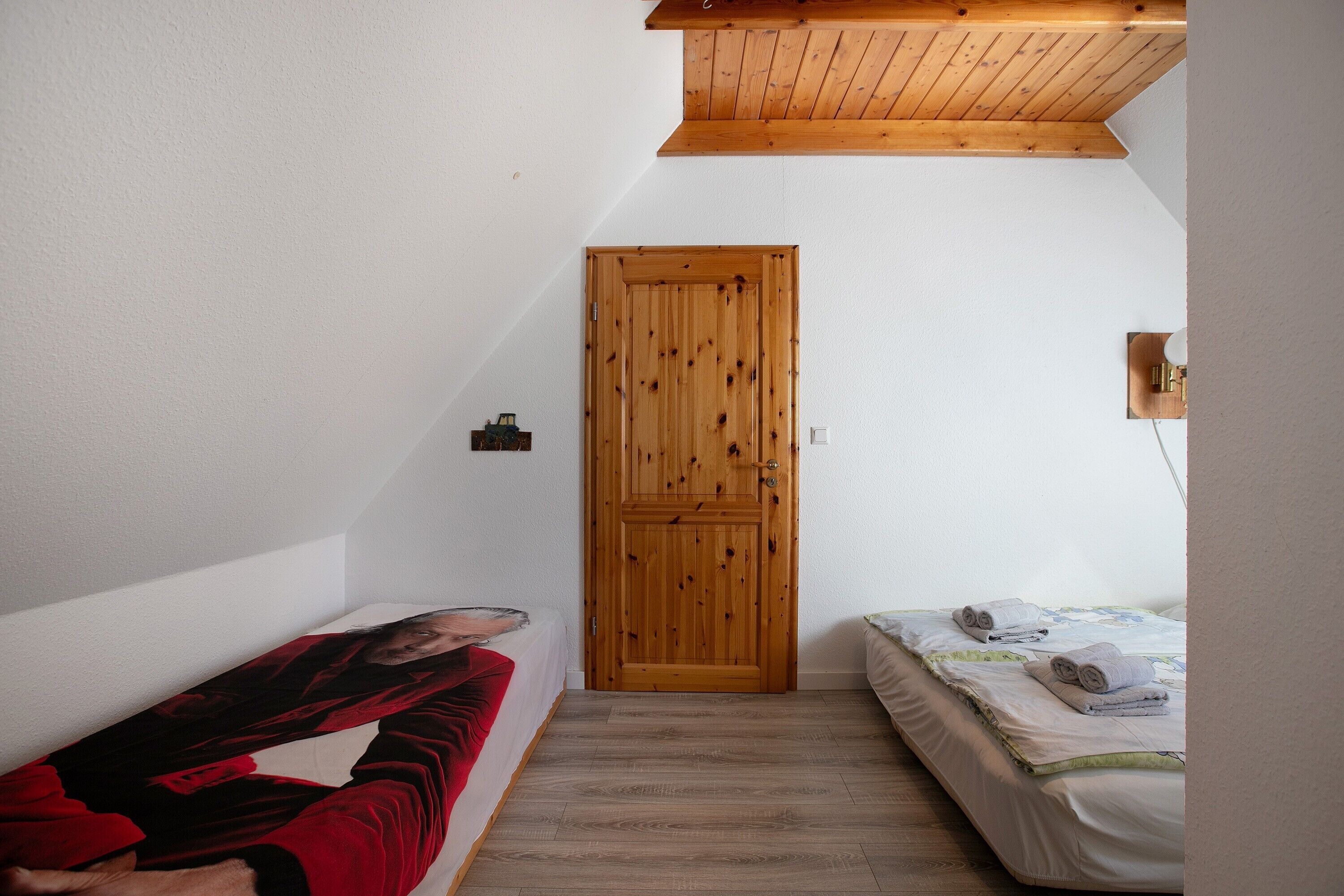 Une chambre avec une porte en bois, un lit avec un matelas blanc et une couverture verte, et une couverture rouge sur le sol.