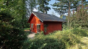 Une cabane rouge au toit en pente, entourée d’une végétation luxuriante et d’arbres.