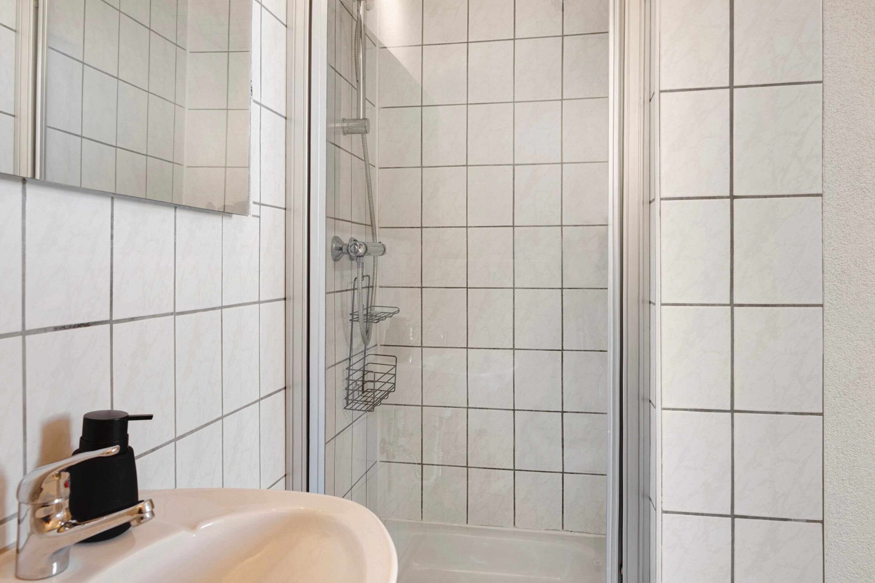 Baño con lavamanos, espejo, ducha y paredes con azulejos.