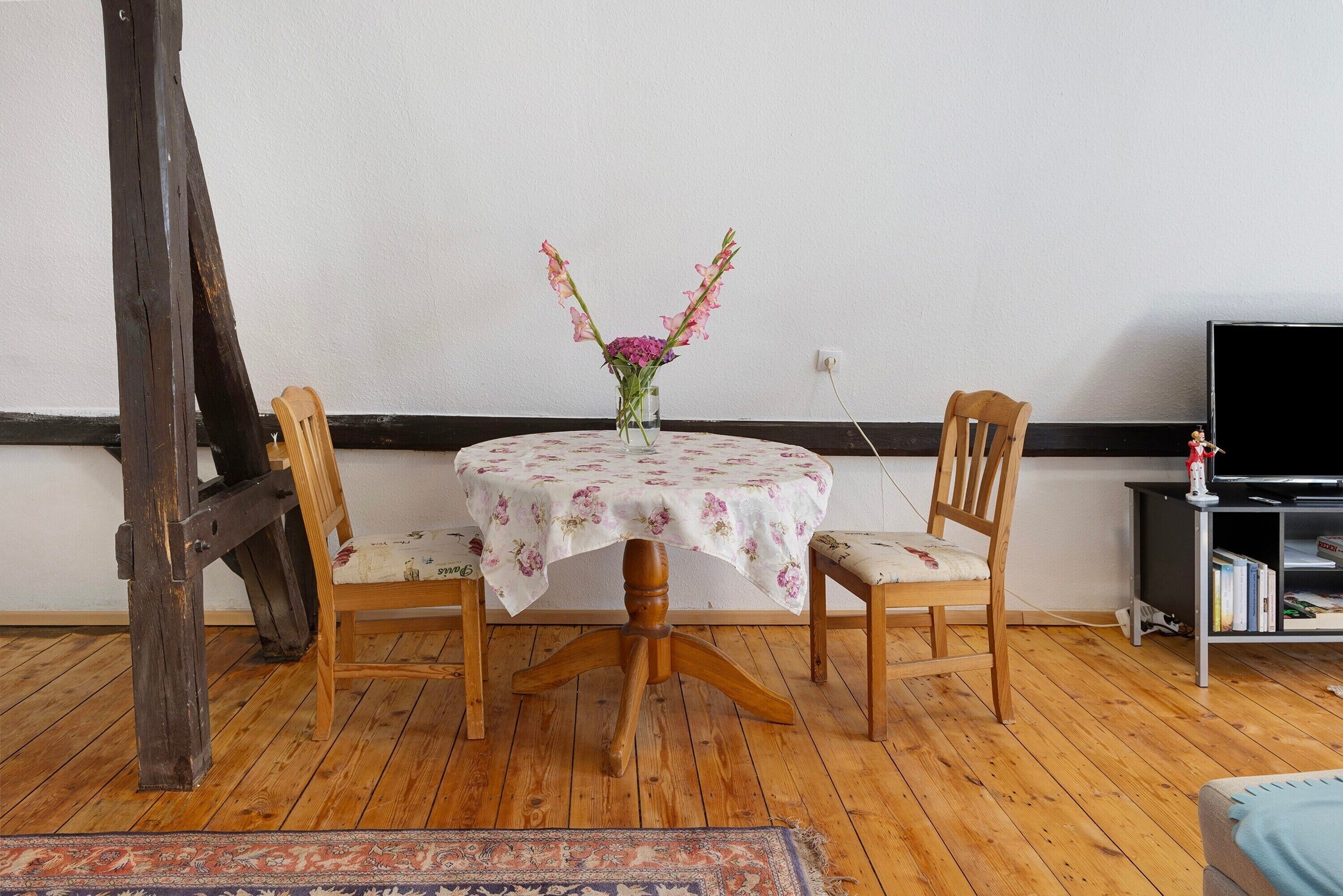 Ein Esstisch mit einer blumigen Tischdecke, zwei Holzstühlen und einer Blumenvase.