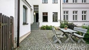 Une cour avec une table et un banc en bois, une clôture en bois et un bâtiment doté de nombreuses fenêtres et d’un escalier.