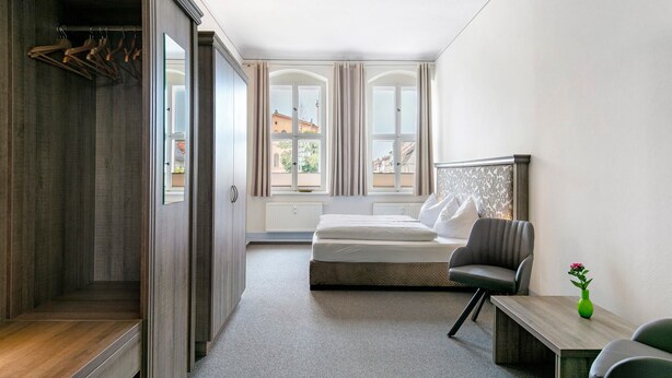 Een slaapkamer met een bed, een stoel, een klein tafeltje, een groene vaas en een uitzicht op gebouwen door het raam.