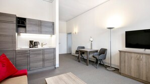 Een moderne keuken met grijze kastjes, een koffiezetapparaat en een spoelbak. Er is een zithoek met een tafel en stoelen, een televisie aan de muur en een rode kussen op de bank.