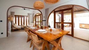 Un comedor con una mesa de madera dispuesta para cuatro personas, rodeada de sillas tejidas. Dos lámparas de mimbre cuelgan sobre la mesa.