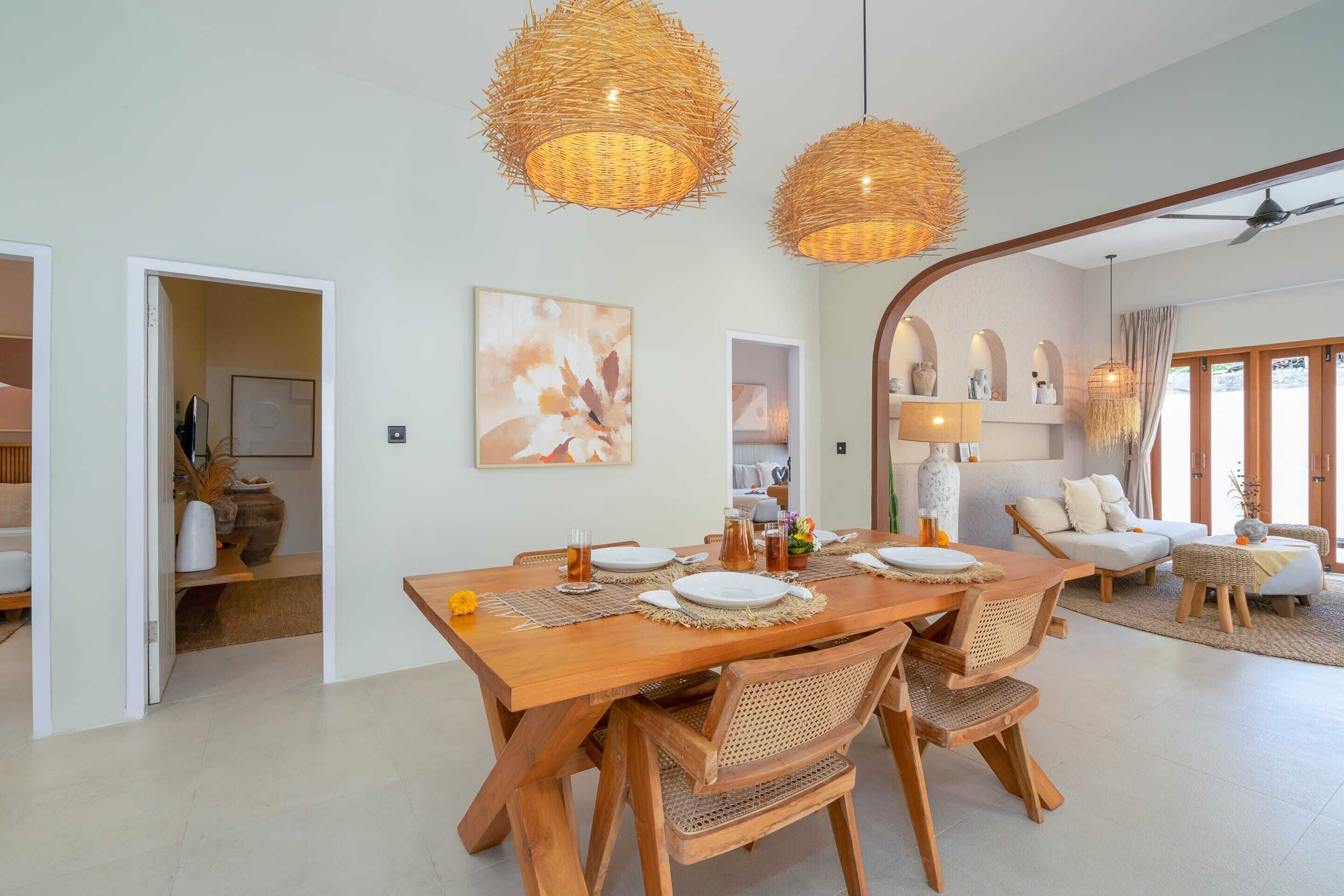 Un comedor con una mesa de madera dispuesta para cuatro personas, rodeada de sillas de mimbre. Dos grandes lámparas colgantes de tejido iluminan la mesa.