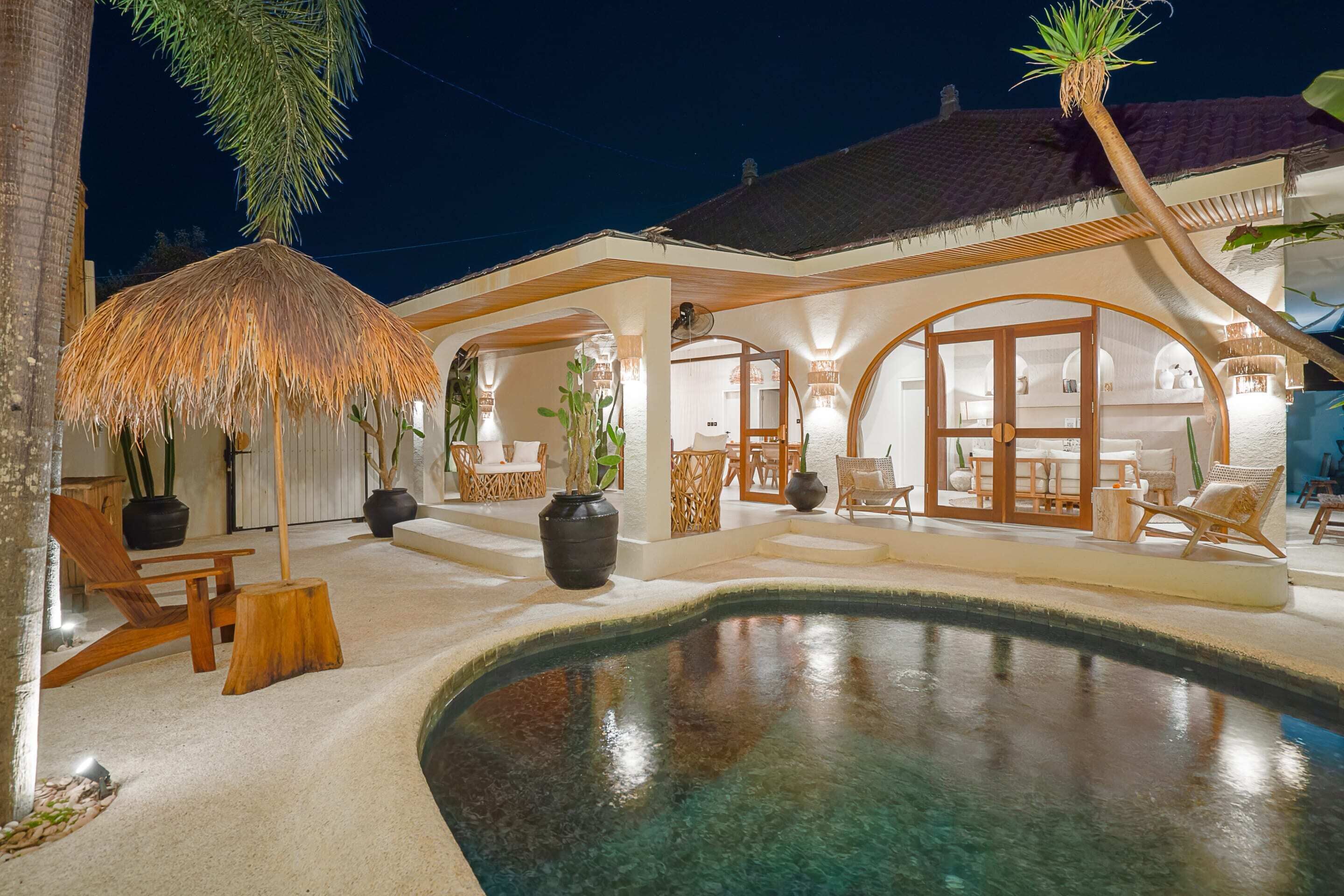 A well-lit outdoor area with a pool, wooden furniture, and a thatched umbrella.
