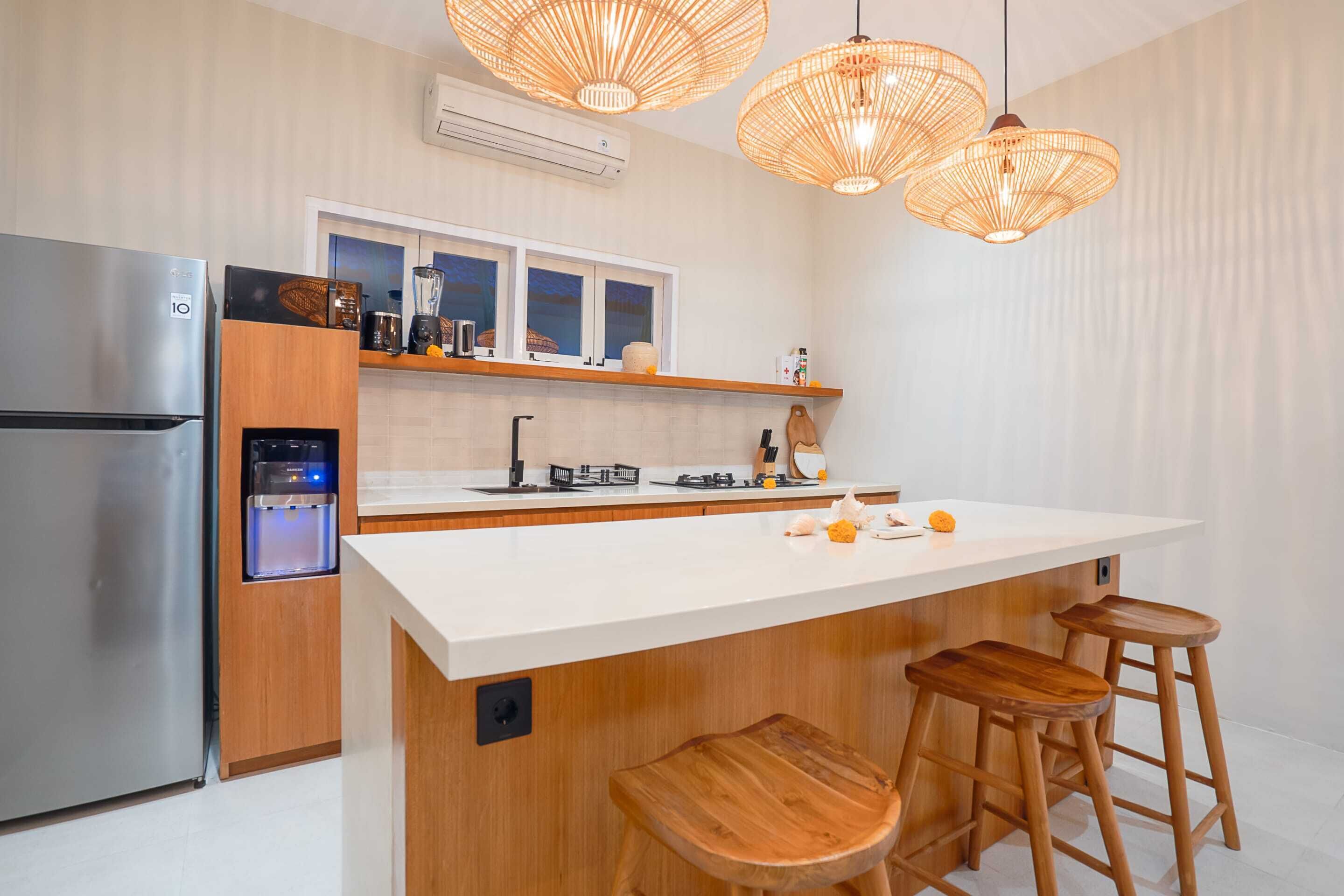 A modern kitchen with a stainless steel refrigerator, a water dispenser, a white countertop, wooden stools, and wicker pendant lights.