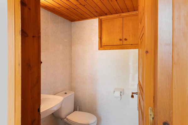 Un baño pequeño con techo de madera, inodoro blanco y un mueble de madera sobre el lavamanos.