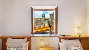 Una habitación con un banco de madera, una mesa pequeña, un ventanal con una persiana abierta y vistas a un edificio de piedra.