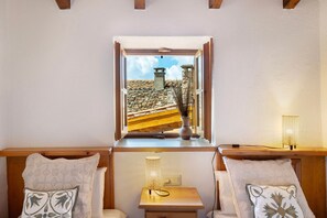 A room with a wooden bench, a small table, a window with an open shutter, and a view of a stone building.