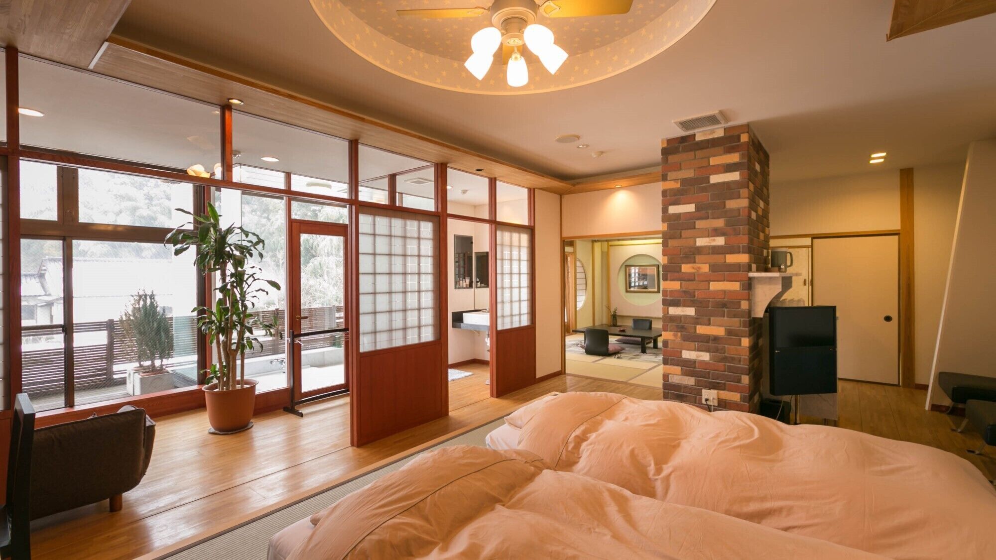 A bedroom with a large bed, sliding glass doors, a potted plant, and a brick fireplace.