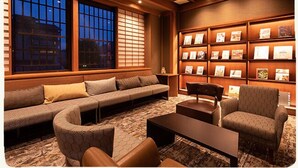 Ein modernes Wohnzimmer mit einer Couch, Sesseln und einem Couchtisch. Ein Bücherregal mit gerahmten Bildern und ein Fenster mit Jalousien.