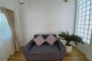 A grey sofa with two cushions, a potted plant, and a window with a grid pattern. - Zenzen Pool Villa (San Sai)