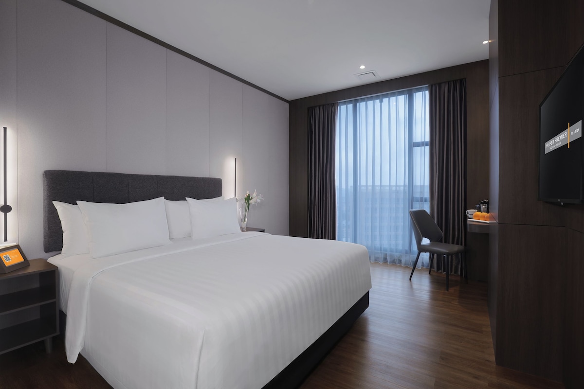 A modern hotel room with a large bed, a desk with a chair, a flat-screen TV, and a large window with curtains.