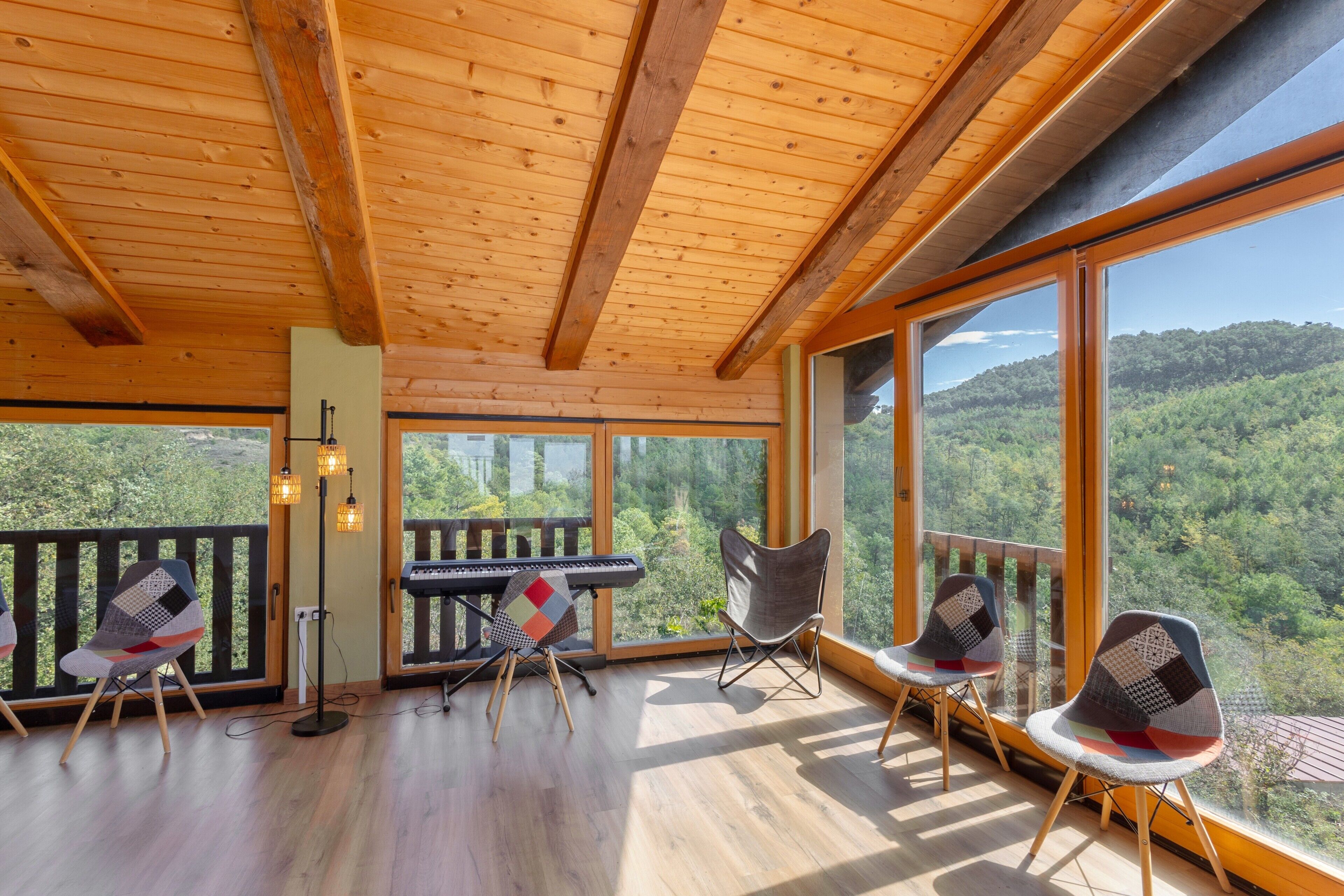 A spacious room with a wooden ceiling, large windows offering a view of a forested landscape, a piano, and modern chairs.