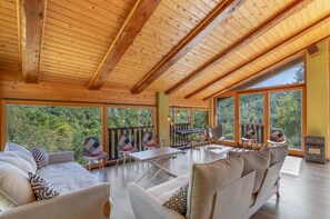 Ein geräumiger Raum mit hoher Decke, großen Fenstern und Blick auf einen Wald. Ein Tisch mit Stühlen und einer Tastatur ist vorhanden.