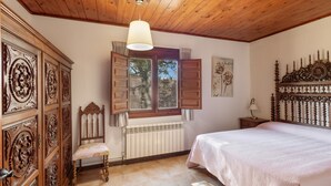 A bedroom with a wooden ceiling, a bed with a white bedspread, a wooden nightstand, a chair, a window with shutters, and a radiator. - Chalet 'Masia La Clariana' with Mountain Views, Private Pool and Wi-Fi (Castelltallat)
