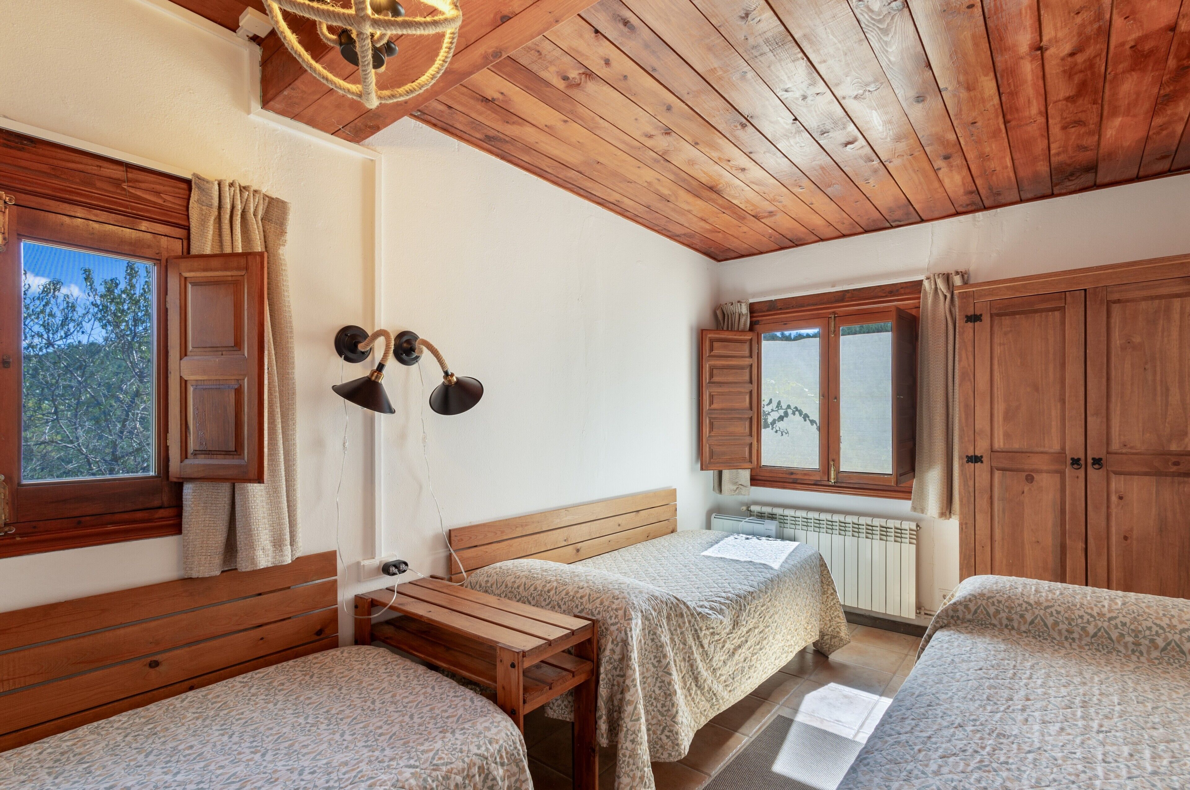 A room with wooden ceiling and walls, a bed with a wooden headboard, a bench, a small table, and a window with curtains.