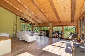 A spacious living area with a wooden ceiling, large windows, a sofa, chairs, and a keyboard.