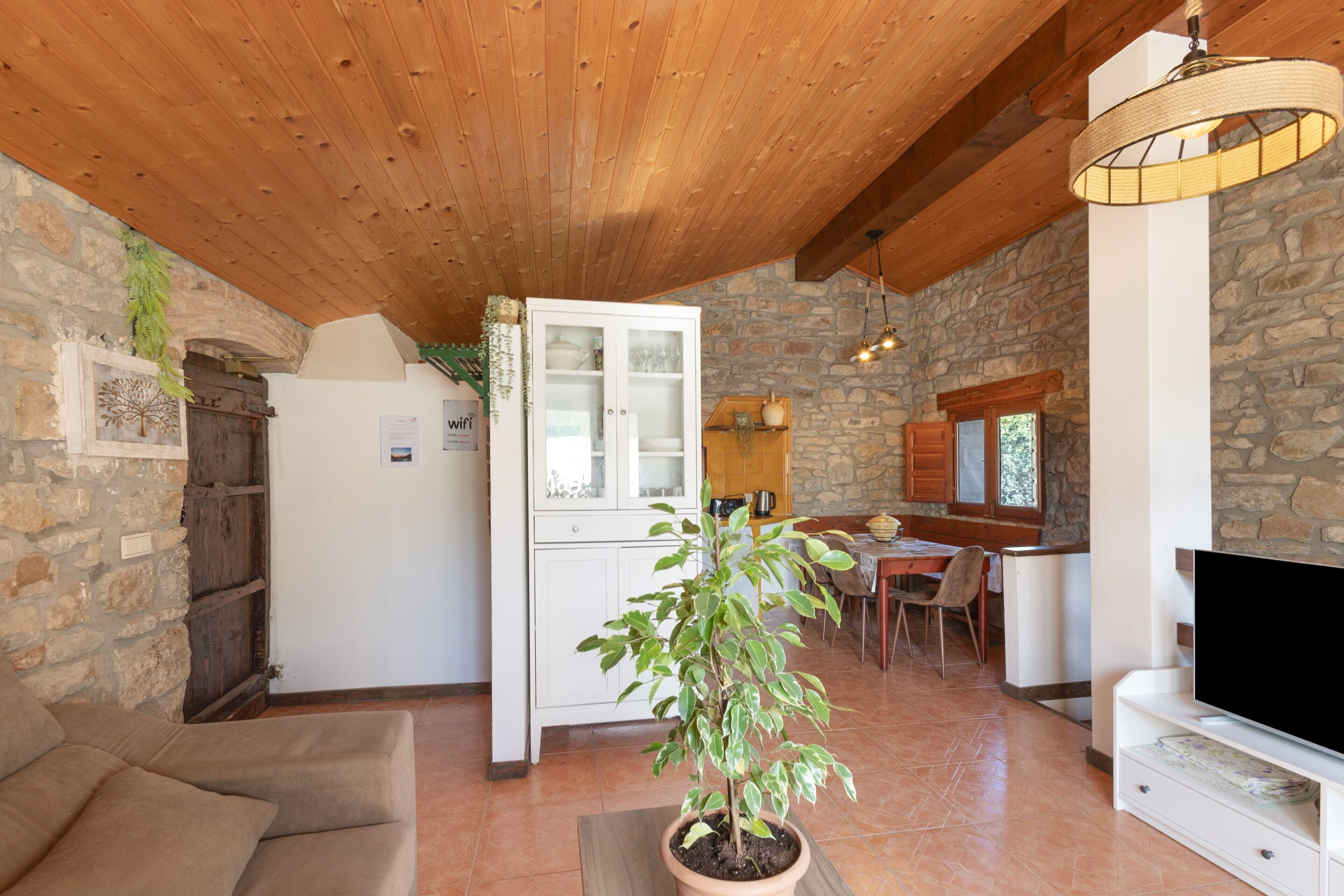 A cozy living area with a wooden ceiling, a stone wall, a dining table with chairs, a television, and a potted plant.