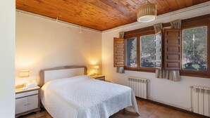 A bedroom with a wooden ceiling, a bed with a white bedspread, a nightstand with a lamp, a window with shutters, and a radiator. - Chalet 'Masia La Clariana' with Mountain Views, Private Pool and Wi-Fi (Castelltallat)