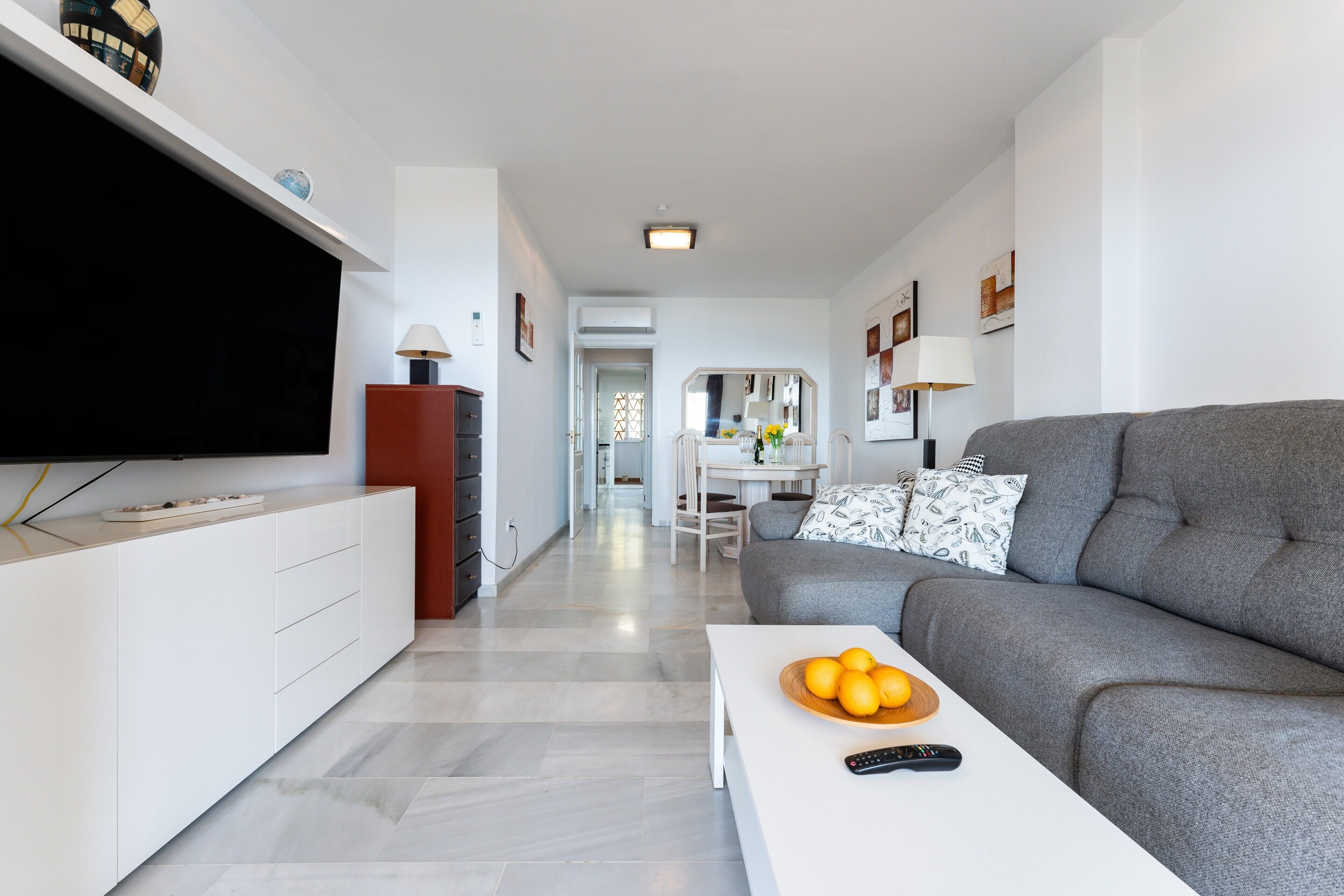 Un salon moderne avec un téléviseur à écran plat, un canapé gris, une table basse ornée d’oranges et un meuble blanc.