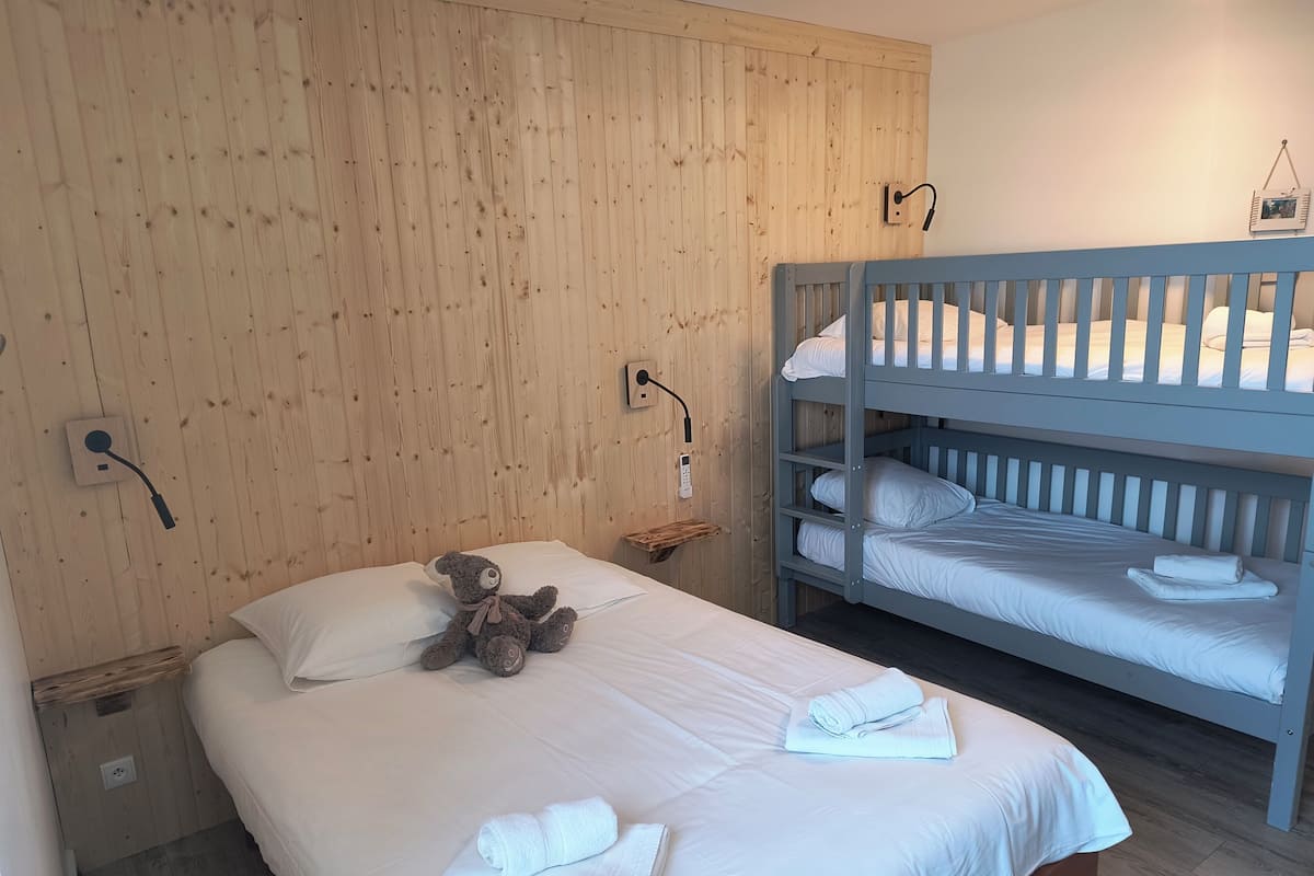 A bedroom with a wooden wall, bunk beds, a bedside table with a lamp, a teddy bear, and neatly folded towels.