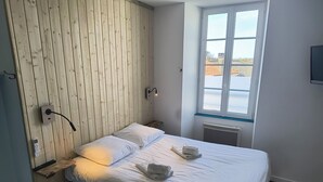A bedroom with a wooden headboard, a bed with white linens, a bedside table with a lamp, and a window with a view of buildings. - Hôtel Bar Restaurant La Marie Galante (Saint-Georges-d'Oleron)