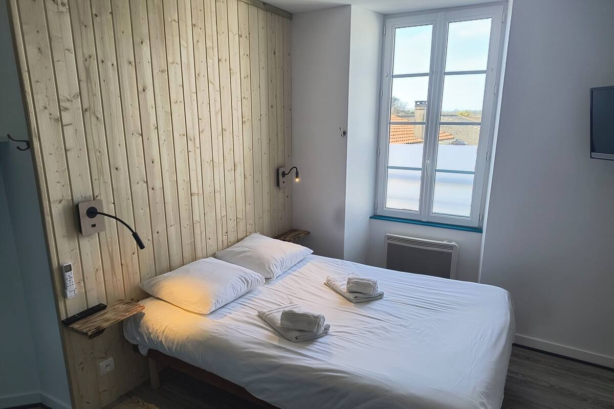 A bedroom with a wooden headboard, a bed with white linens, a bedside table with a lamp, and a window with a view of buildings.