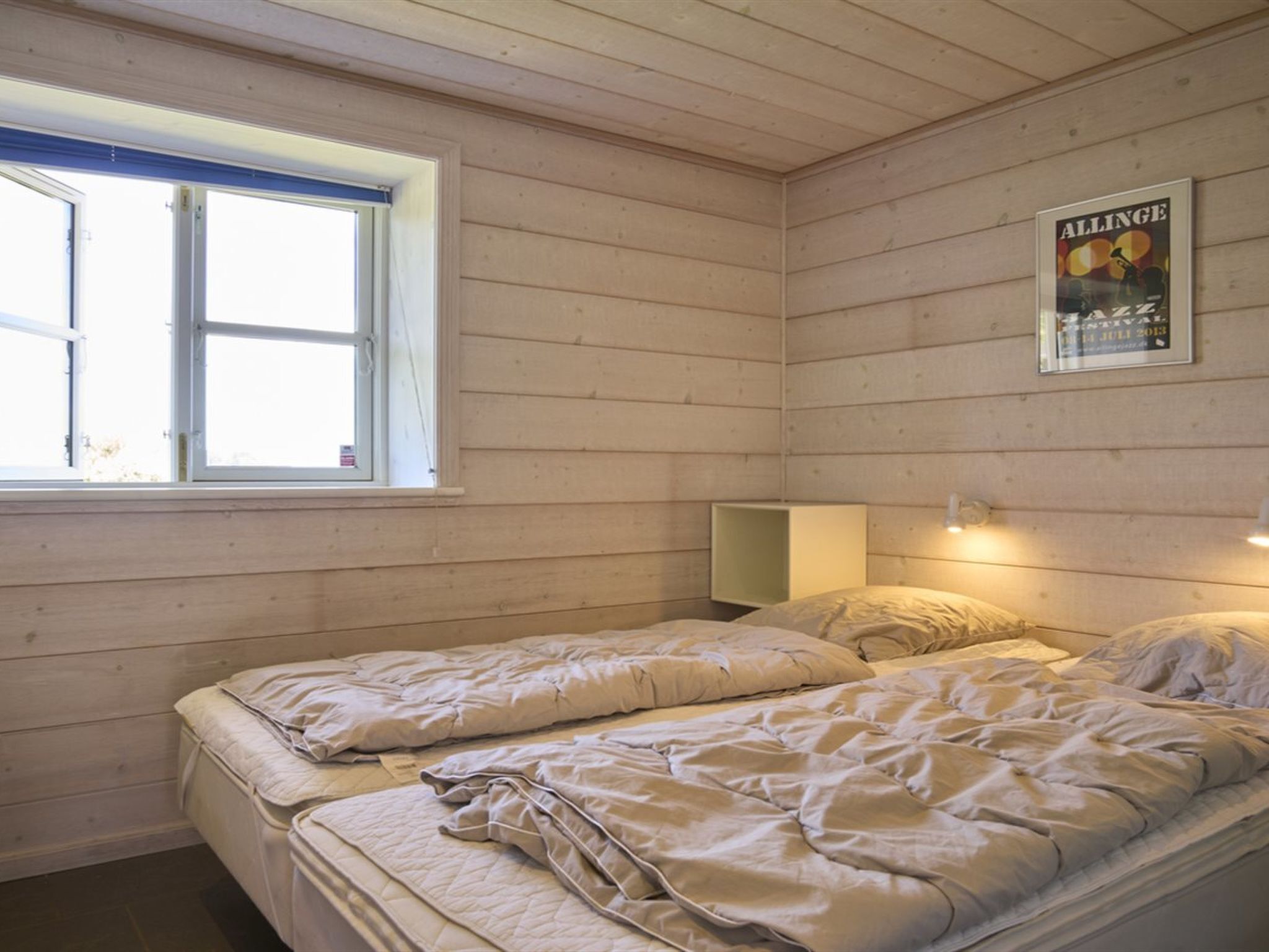 Een kamer met twee bedden, een raam, een poster aan de muur en een nachtkastje met een lamp.