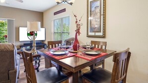 Un coin repas avec une table en bois dressée pour quatre personnes, entourée de chaises. Un téléviseur est fixé au mur, et il y a une fenêtre avec des stores.