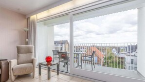 Varanda com vista para casas, uma cadeira, uma mesa pequena com um vaso vermelho e um abajur.
