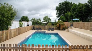 A rectangular swimming pool with a wooden fence surrounding it, a covered seating area with chairs, and a palm tree in the background.