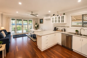 A modern kitchen with white cabinetry, a built-in dishwasher, and a dining area with a table and chairs. - Sea Renity On Earnshaw (West Busselton)