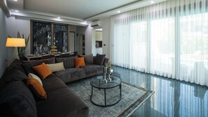 A modern living room with a dark-colored sofa, a glass coffee table, and a large window with sheer curtains. - Living Dream Villa Harmony (Antalya)