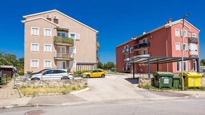 A residential area with two multi-story apartment buildings, a parking area with cars, and green and yellow waste bins. - Apartment White (Rovinj)