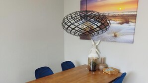 A dining area with a wooden table, blue chairs, a hanging pendant light, and a painting of a sunset on the wall.