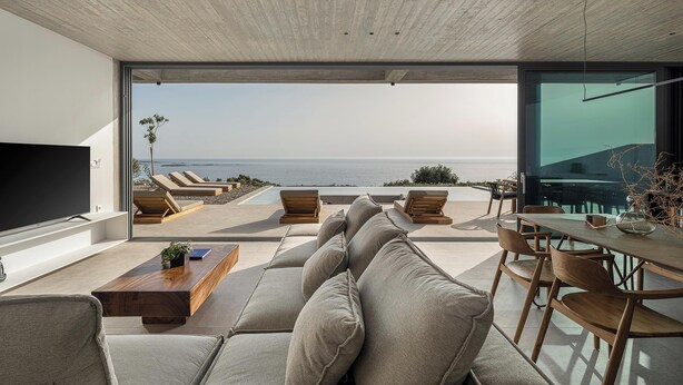 Een moderne woonkamer met een grote bank, een salontafel en een eethoek met stoelen. Links bevindt zich een televisie en er is uitzicht op de oceaan door grote ramen.